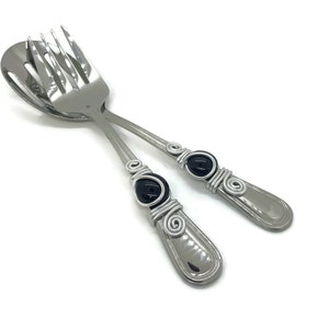 May include: A pair of stainless steel salad servers. Each server has a handle with a black stone accent wrapped in silver-colored wire. The spoon and fork are angled across each other against a white background.