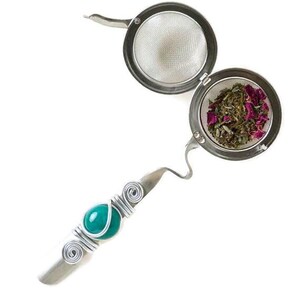 May include: A silver tea infuser with a teal blue gemstone accent. The infuser has a round, mesh basket for loose tea leaves. The basket is open, revealing dried herbs and rose petals.