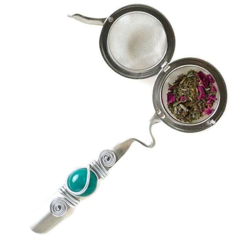 May include: A silver tea infuser with a teal blue gemstone accent. The infuser has a round, mesh basket for loose tea leaves. The basket is open, revealing dried herbs and rose petals.