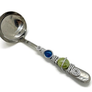 Può includere: Un mestolo d'argento con un manico di perline di vetro blu e verde. Il manico è avvolto in filo e le perline sono fissate con filo.