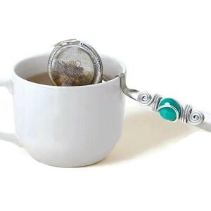 May include: A silver tea infuser with a turquoise bead on the handle, sitting in a white teacup.