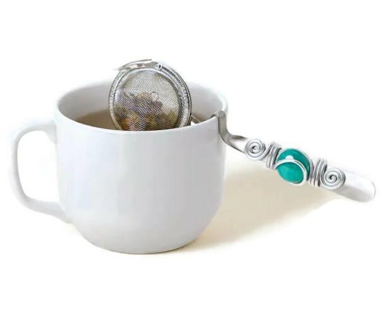 May include: A silver tea infuser with a turquoise bead on the handle, sitting in a white teacup.