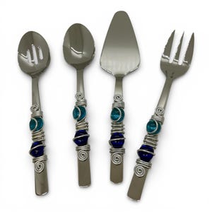 Op de afbeelding: Een set van vier roestvrijstalen serveerbestekken. Elk bestek heeft een decoratief handvat met blauwe en turquoise glazen kralen gewikkeld in zilverkleurig draad. De set bevat een sleef, lepel, taartschep en vork.