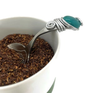 May include: A silver tea infuser spoon with a teal blue glass bead on the handle. The spoon is resting in a white mug filled with loose leaf tea.