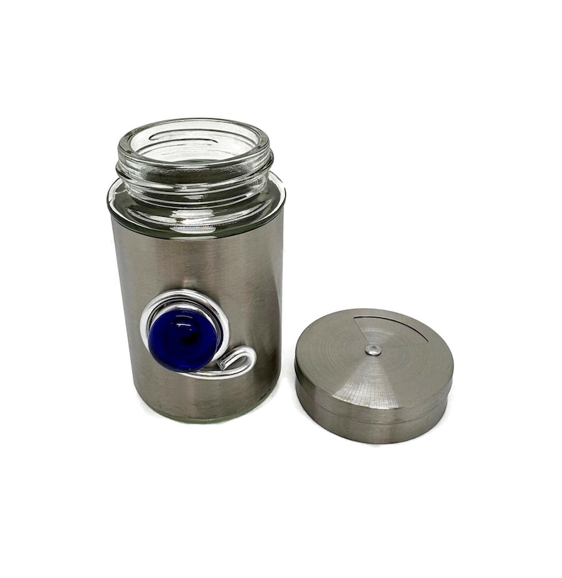 May include: A silver spice jar with a blue glass eye charm. The jar has a clear glass top and a silver lid.