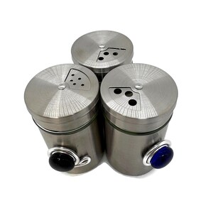 May include: Set of three stainless steel spice shakers with glass jars and colored glass knobs. The shakers have a hole pattern on the top for dispensing spices.