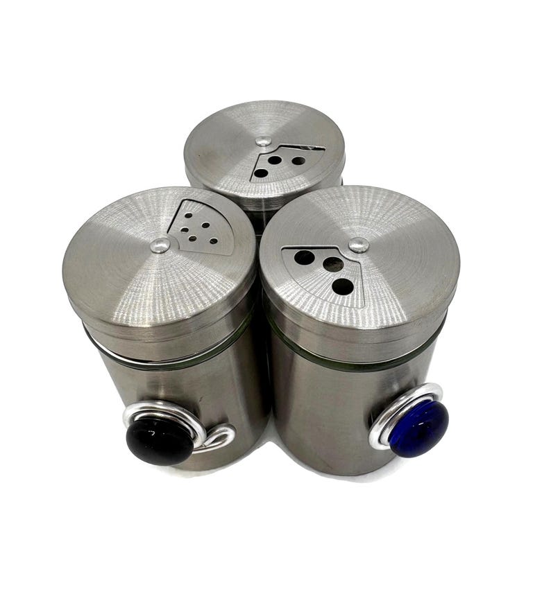 May include: Set of three stainless steel spice shakers with glass jars and colored glass knobs. The shakers have a hole pattern on the top for dispensing spices.
