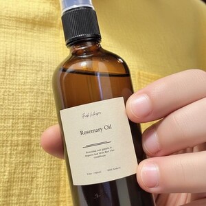 May include: A brown glass bottle of Rosemary Oil with a black and clear spray top. The label reads "Rosemary Oil" with additional text. The bottle is held in a person's hand against a yellow background.