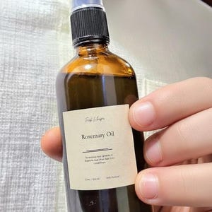May include: A brown glass bottle of Rosemary Oil with a black and clear spray top. The label reads "Rosemary Oil" with additional text. The bottle is held in a person's hand.