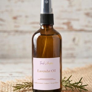 Könnte beinhalten: Eine braune Glassprühflasche mit Lavendelöl, schwarzem Verschluss und weißem Etikett. Auf dem Etikett steht "Fresh Whispers Lavendar Oil" mit dem Text "Kaltgepresst | Natürlich | Ohne Zusatzstoffe". Die Flasche steht auf einer Jutefläche.
