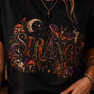 May include: A black t-shirt featuring a colourful graphic print with the word STRANGE in stylised lettering. The design includes a black cat, a moth, mushrooms, a crescent moon, a skull, and a candle, all surrounded by various botanical elements.