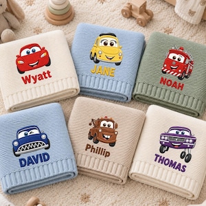 May include: Six folded knit blankets featuring embroidered cartoon vehicles and names. The blankets are cream with a red car labelled Wyatt, blue with a yellow car labelled JANE, green with a red fire truck labelled NOAH, light blue with a blue car labelled DAVID, tan with a brown truck labelled Phillip, and cream with a purple car labelled THOMAS.
