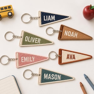 Könnte beinhalten: Sechs dreieckige Wimpel-Schlüsselanhänger aus Filz mit Metallringen, jeweils mit gestickten Namen: Liam in Marineblau, Noah in Hellbraun, Oliver in Olivgrün, Emily in Rosa, Ava in Creme und Braun sowie Mason in Petrol, neben einem Spielzeugbus und Apfel.