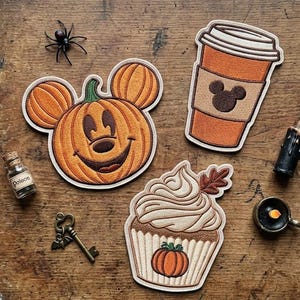 May include: Three embroidered patches featuring a pumpkin with mouse ears, a coffee cup with a mouse icon, and a cupcake with a small pumpkin. The scene includes a small glass bottle, a metal key, a black candle, and a small black spider.