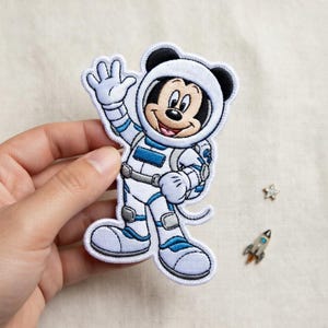 Könnte beinhalten: Gestickter Aufnäher mit Mickey Mouse in einem weißen und blauen Astronautenanzug, der winkt. Der Aufnäher hat einen weißen Rand und wird von einer Hand gehalten. Ein kleiner Stern und ein raketenförmiger Anhänger befinden sich im Hintergrund.
