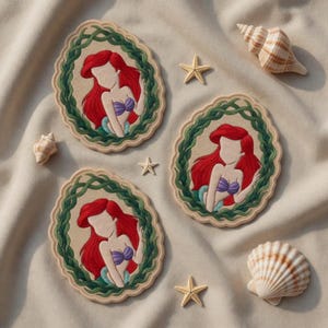 May include: Three embroidered coasters featuring a red-haired mermaid with a purple seashell top, surrounded by a green vine border. The coasters are oval-shaped and set on a beige surface with seashells and starfish.