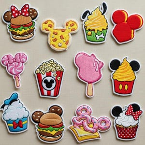 May include: A collection of embroidered patches featuring various food items and Mickey Mouse-inspired designs. Includes a burger, pizza, cupcake, popcorn, ice cream, and donuts. The patches are colorful with detailed stitching and white borders.