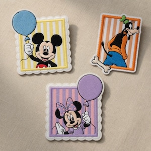 Op de afbeelding: Drie geborduurde patches met Disney-personages. Mickey Mouse houdt een blauwe ballon vast tegen een geel gestreepte achtergrond. Goofy staat op een bot, tegen een oranje en wit gestreepte achtergrond. Minnie Mouse houdt een paarse ballon vast tegen een roze gestreepte achtergrond.