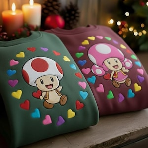 May include: Two sweatshirts featuring cartoon characters. One is forest green with a mushroom character and the other is burgundy with a female mushroom character. Both have colorful heart accents and are surrounded by a heart border.