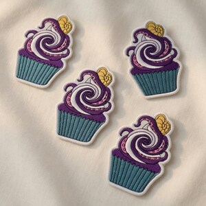 Può includere: Quattro toppe ricamate a forma di cupcake. Ogni cupcake ha una base blu-azzurro, glassa viola, vortici bianchi, accenti rosa e un disegno giallo a forma di conchiglia. Sfondo bianco.