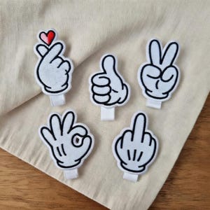 Puede incluir: Cinco parches blancos de manos de dibujos animados con contornos negros que representan diferentes gestos: un corazón, un pulgar hacia arriba, el signo de la paz, el signo de ok y el dedo corazón. Cada parche tiene una cinta blanca.