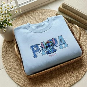 Puede incluir: Sudadera azul claro con un dise&ntilde;o de personaje de dibujos animados y el nombre "Jason" debajo. El dise&ntilde;o incluye la palabra "PAPA" en una fuente universitaria, con el personaje en el centro. La sudadera est&aacute; en una cesta tejida.