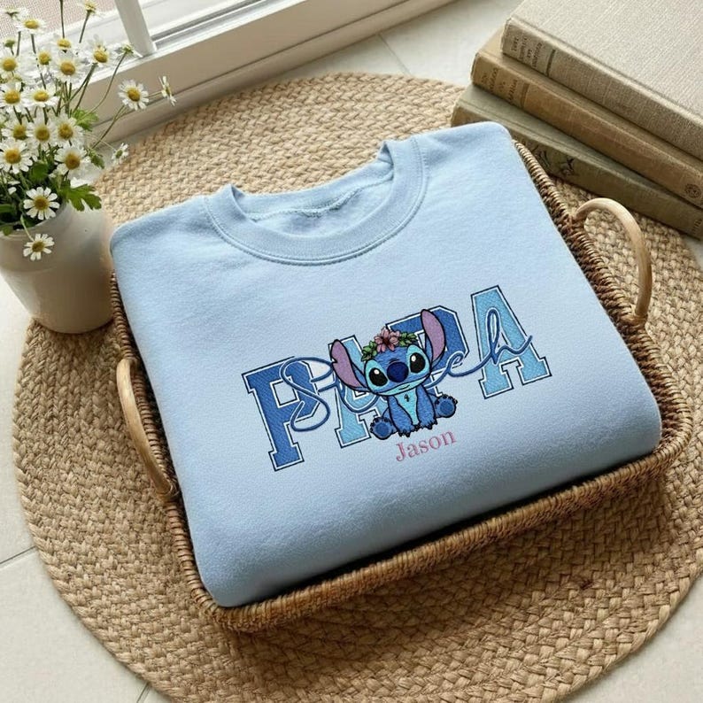 Puede incluir: Sudadera azul claro con un dise&ntilde;o de personaje de dibujos animados y el nombre "Jason" debajo. El dise&ntilde;o incluye la palabra "PAPA" en una fuente universitaria, con el personaje en el centro. La sudadera est&aacute; en una cesta tejida.