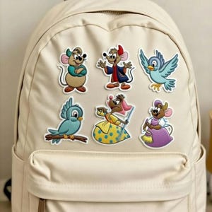 May include: A beige backpack adorned with embroidered patches featuring cartoon mice and birds. The patches showcase characters in various outfits and poses, with vibrant colors like blue, yellow, and purple. The backpack has a simple design with a front pocket.