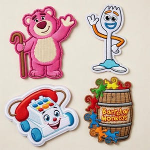 Puede incluir: Cuatro parches bordados con personajes de una popular película de animación. Incluye un oso rosa con un bastón, un tenedor blanco con brazos naranjas, un teléfono de juguete sonriente y un barril de monos con el texto "Barrel of Monkeys".