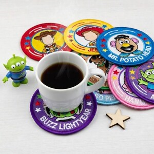 May include: A white coffee mug filled with dark liquid sits on a purple coaster with "BUZZ LIGHTYEAR" text. Several colorful, round coasters with cartoon character designs surround the mug. A small green alien figurine and a wooden star are also present.