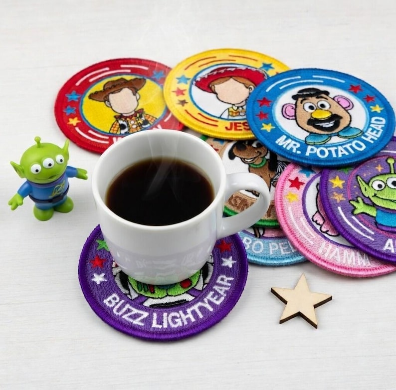 May include: A white coffee mug filled with dark liquid sits on a purple coaster with "BUZZ LIGHTYEAR" text. Several colorful, round coasters with cartoon character designs surround the mug. A small green alien figurine and a wooden star are also present.