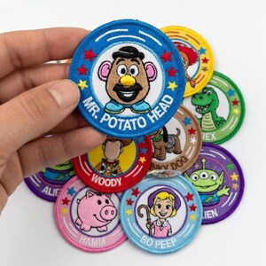 May include: A collection of embroidered patches featuring characters from the animated film Toy Story. The patches are circular, with blue, red, green, and purple borders. Characters include Mr. Potato Head, Woody, and Bo Peep.