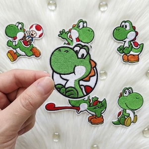 May include: A collection of embroidered fabric patches featuring the green dinosaur character Yoshi in various poses, including one patch showing Yoshi with a red and white spotted mushroom character, all with detailed stitching and vibrant colours.