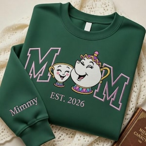May include: A dark green sweatshirt with the word "MOM" in pink block letters, along with a cartoon teacup and teapot. The sleeve reads "Mimmy" in pink script, and the year "EST. 2026" is below the cartoon characters.