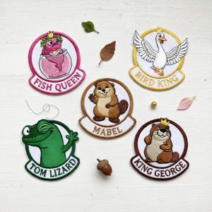 May include: Embroidered patches featuring cartoon characters: a fish in a bowl labeled "Fish Queen", a goose labeled "Bird King", a beaver named "Mabel", a lizard labeled "Tom Lizard", and a beaver labeled "King George". Each patch has a unique design and color scheme.