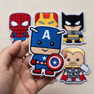 May include: A collection of embroidered fabric patches featuring stylized cartoon superheroes. The foreground patch depicts Captain America in blue, red, and white with a shield. Other patches show Spider-Man, Iron Man, Batman, and Thor designs.