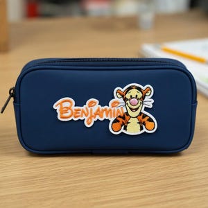 Könnte beinhalten: Ein rechteckiges, marineblaues Federmäppchen mit schwarzem Reißverschluss. Auf der Vorderseite befindet sich ein gestickter Aufnäher der Figur Tigger sowie der Name Benjamin in orangefarbener Schreibschrift mit weißem Rand.