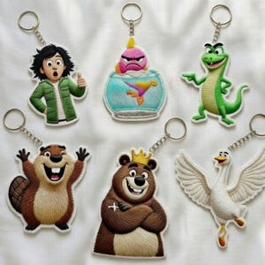 May include: A collection of cartoon character keychains. The keychains feature various designs, including a boy, a fish in a bowl, a green lizard, a beaver, a bear with a crown, and a white swan. Each keychain has a silver-colored ring.