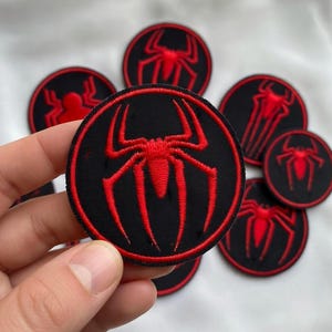 May include: A circular embroidered patch featuring a vibrant red spider emblem centered on a black background, enclosed by a thin red circular border. The design is stitched with dense thread, creating a textured surface on the round fabric patch.