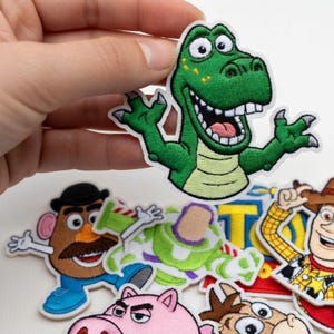 May include: Embroidered patches featuring characters from a popular animated film. A green dinosaur patch is held up, with other character patches visible below. The patches have white borders and are colorful.