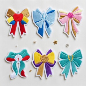 May include: Six embroidered bow patches in various colours, including red, blue, pink, yellow, and teal. Each patch features a unique design, such as a rose or a jewel, with white stitching outlining the bows. The patches are arranged on a white surface.