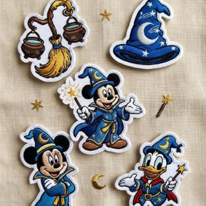 Puede incluir: Parches bordados con diseños de magos. Incluyen a Mickey Mouse, Minnie Mouse y el Pato Donald vestidos de magos, un sombrero de mago, una escoba con calderos y detalles de estrellas. Los parches son en tonos azules, dorados y blancos.