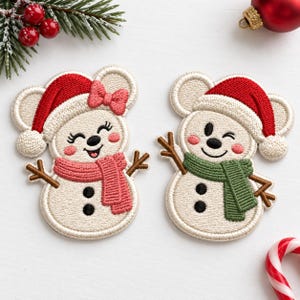 May include: Two embroidered snowman patches featuring white bodies, black button details, and brown stick arms. One snowman wears a red hat with a pink bow and a pink scarf, while the other wears a red hat and a green scarf. Both have rosy cheeks.