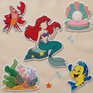 Puede incluir: Un conjunto de parches bordados que incluye un cangrejo rojo, una sirena de pelo largo y rojo sobre una roca, una concha rosa y verde azulado con una perla, un pez amarillo y azul, y un grupo de corales verdes, rosas y morados con burbujas.