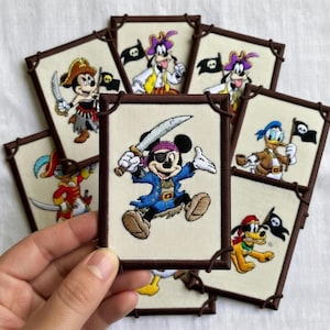 May include: Embroidered patches featuring Disney characters dressed as pirates. Mickey Mouse, Goofy, Donald Duck, and Pluto are depicted with swords and pirate flags. Each patch has a brown border and is on a cream-colored background.