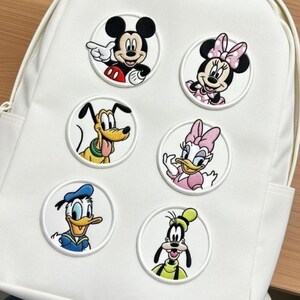 May include: White backpack decorated with six embroidered patches featuring Disney characters. The patches include Mickey Mouse, Minnie Mouse, Pluto, Daisy Duck, Donald Duck, and Goofy. The patches are round with white borders and colorful character designs.