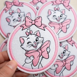 May include: Embroidered patches featuring a white cat with a pink bow and a wink. The cat is inside a white circle with a pink border, and a pink bow is at the top. The patches are on a white background.