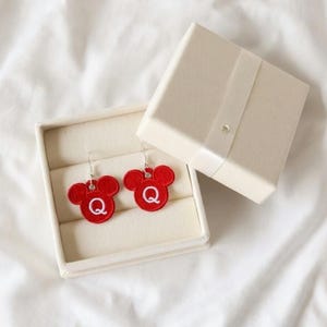 Pode incluir: Brincos vermelhos em forma de Mickey Mouse com a letra "Q" bordada em branco. Apresentados numa caixa de joias creme com uma fita a condizer e um pequeno cristal.