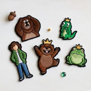 May include: Embroidered patches featuring cartoon characters, including a bear, a frog, a lizard, and a person, all with crowns. The patches are in various colors, including brown, green, and blue. An acorn and a green gem are also present.