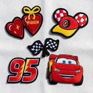 Puede incluir: Un conjunto de parches bordados con diseños de la película "Cars". Incluye un corazón rojo con el texto "McQueen", una gorra de carreras, banderas a cuadros, el número "95" con un rayo y un coche de carreras rojo.